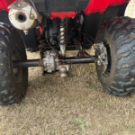 Underside of a red ATV showing large off-road tires, suspension, drivetrain, and hitch on dry grass surface.