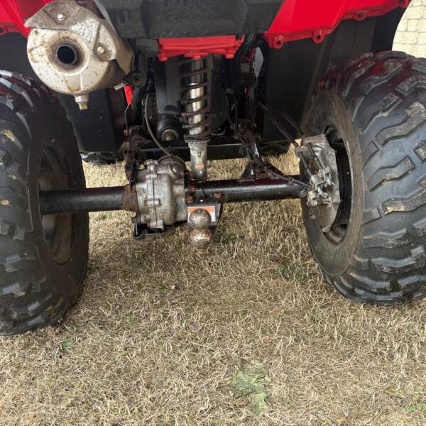 Underside of a red ATV showing large off-road tires, suspension, drivetrain, and hitch on dry grass surface.