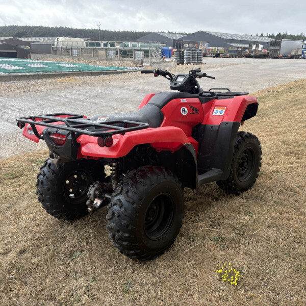 Red Honda ATV parked on dry grass at a construction site with warehouses and a blue truck in the background.
