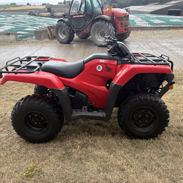 Red Honda four-wheeled ATV parked on muddy grass with farm buildings in the background and a tractor nearby.