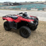 Red all-terrain vehicle (ATV) parked on dry grass with a blue-green tarp-covered roof and industrial buildings in the background.
