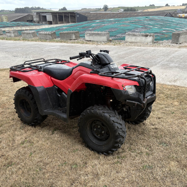 Red all-terrain vehicle (ATV) parked on dry grass with a blue-green tarp-covered roof and industrial buildings in the background.