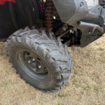 Close-up of a muddy off-road vehicle wheel and suspension showing large tread tire and rusted components.