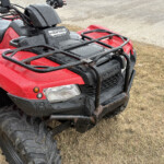 Red Honda FourTrax ATV front quarter view with a cargo rack, wet from rain, on a grassy dirt lot