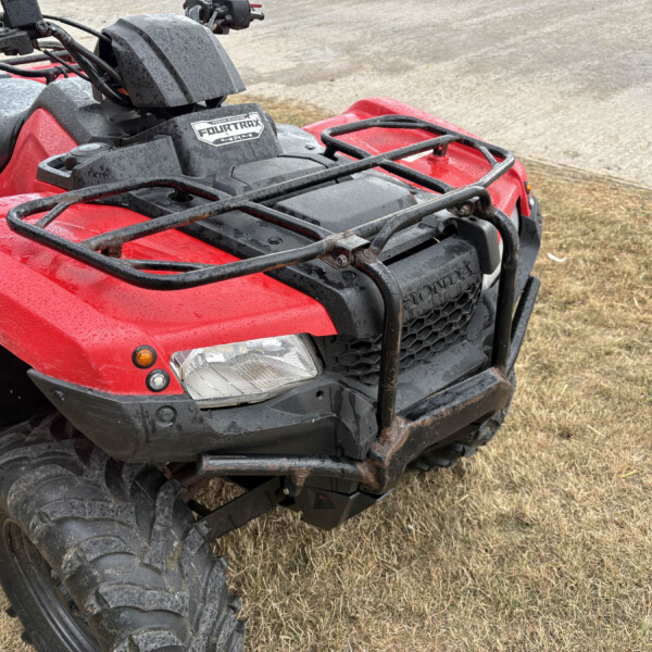 Red Honda FourTrax ATV front quarter view with a cargo rack, wet from rain, on a grassy dirt lot