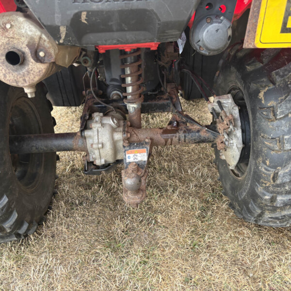 Rear view of a rusted tractor hitch and driveline with large treaded tires on a dry field.