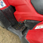 Front-left close-up of a red vehicle body with a warning sticker, mud-splattered black fender, and a textured step plate; dry grass visible nearby.