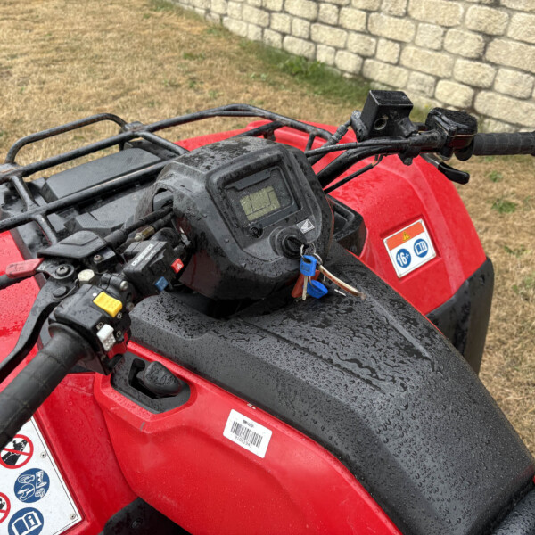 Red ATV handlebars with a wet dashboard, ignition keys, and warning stickers visible beside a stone wall fence behind.