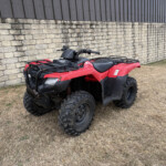 Red all-terrain vehicle (ATV) parked beside a stone wall and industrial building on dry, brown grass.