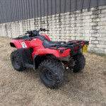 Red quad bike/ATV parked on dry grass beside a stone wall and metal building, license plate visible (UK plate).