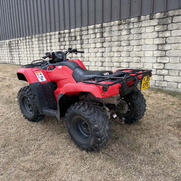 Red quad bike/ATV parked on dry grass beside a stone wall and metal building, license plate visible (UK plate).