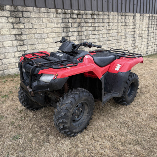 Red all-terrain vehicle (ATV) parked on dry grass beside a stone wall and metal siding building, with a yellow sign to the right