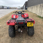 Red ATV foreground in a dry grassy lot, with an industrial yard of trucks and a stone wall on the right under a cloudy sky.