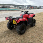 Red ATV parked on a dry grassy area with an industrial yard and trucks in the background.