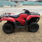 Red ATV parked on dry grass with a construction yard and green tarps behind.