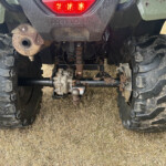 Rear view of a muddy Honda ATV/UTV showing large tread tires, rear suspension, and drive components beneath the bumper light bar location of the exhaust on the left side