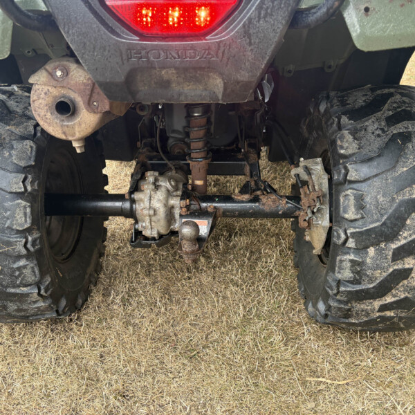Rear view of a muddy Honda ATV/UTV showing large tread tires, rear suspension, and drive components beneath the bumper light bar location of the exhaust on the left side