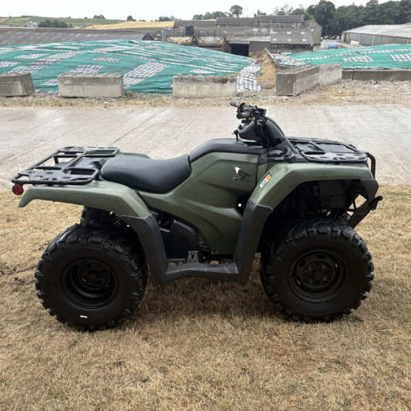 Green ATV parked on dry grass with an industrial yard and tarp-covered slopes in the background.
