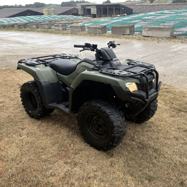 Green all-terrain vehicle parked on dry grass with concrete blocks and a tarp-covered industrial building in the background.