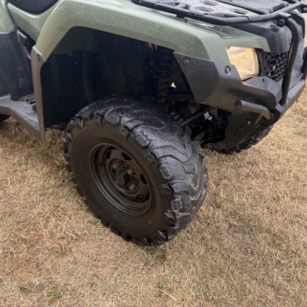 Close-up of an off-road ATV front corner, showing a green body, rugged bumper, headlight, and large muddy tire on grass.
