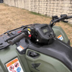 Close-up of a Honda ATV's handlebar and digital display, with warning stickers on the green body and a stone wall in the background.