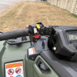Close-up of an ATV handlebar with a yellow toggle switch and black controls, rain droplets on the surface and a warning label on the body.