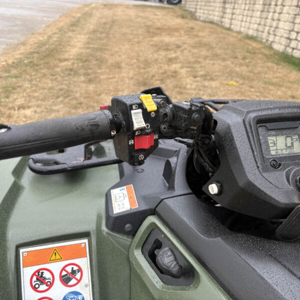 Close-up of an ATV handlebar with a yellow toggle switch and black controls, rain droplets on the surface and a warning label on the body.