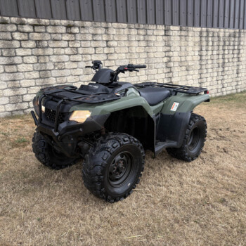 Green ATV parked on dry grass beside a stone wall and metal-clad building, with a yellow sign and traffic cone in the background.