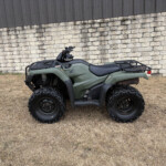 Green Honda ATV with cargo racks parked on dry grass in front of a stone wall and dark wooden fence.