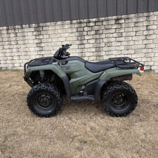 Green Honda ATV with cargo racks parked on dry grass in front of a stone wall and dark wooden fence.