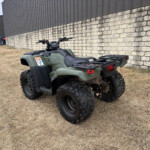 Green Honda ATV parked on dry grass beside a stone retaining wall near an industrial building.