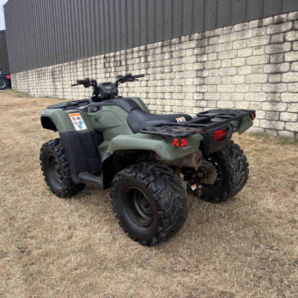 Green Honda ATV parked on dry grass beside a stone retaining wall near an industrial building.