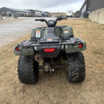Green all-terrain vehicle parked on dry grass in an industrial yard with warehouses and trucks in the background.