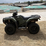 Green Honda ATV parked on dry grass with a construction site and tarped ground in the background.