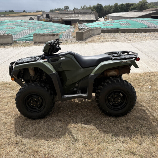 Green Honda ATV parked on dry grass with a construction site and tarped ground in the background.