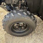 Close-up of an off-road vehicle wheel and chunky tire, with suspension components visible above the tire on dry grass.