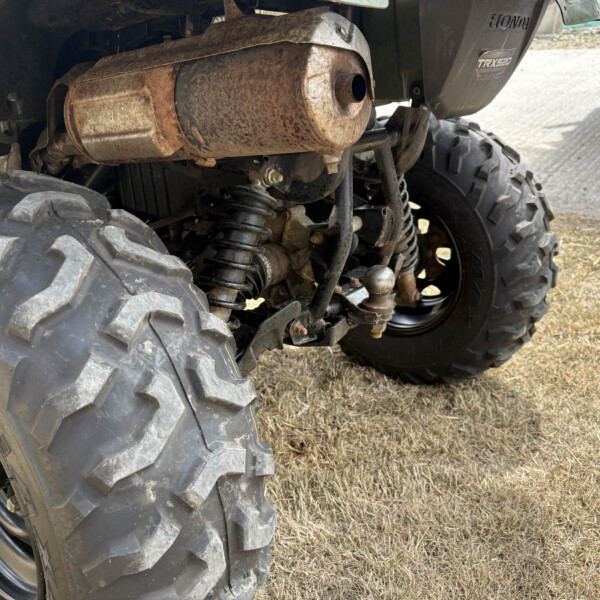 Rear view of a Honda quad bike showing a rusted exhaust canister, suspension, and large knobby tires on dry grass.