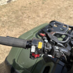 Close-up view of ATV/motorcycle handlebar controls: red kill switch, yellow button, and black switch housing with a green body and dry grass background.