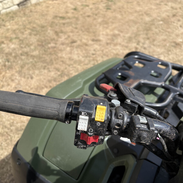 Close-up view of ATV/motorcycle handlebar controls: red kill switch, yellow button, and black switch housing with a green body and dry grass background.