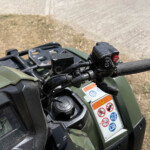 Close-up of an ATV/quad bike handlebar with grip, ignition area, and red 4WD switch visible beside warning labels.