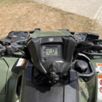 Close-up view of a green Honda ATV handlebar cluster with the speedometer and warning stickers visible below.