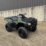 Olive-green ATV parked on dry grass in front of a stone wall and metal siding building