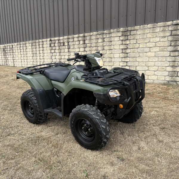 Olive-green ATV parked on dry grass in front of a stone wall and metal siding building