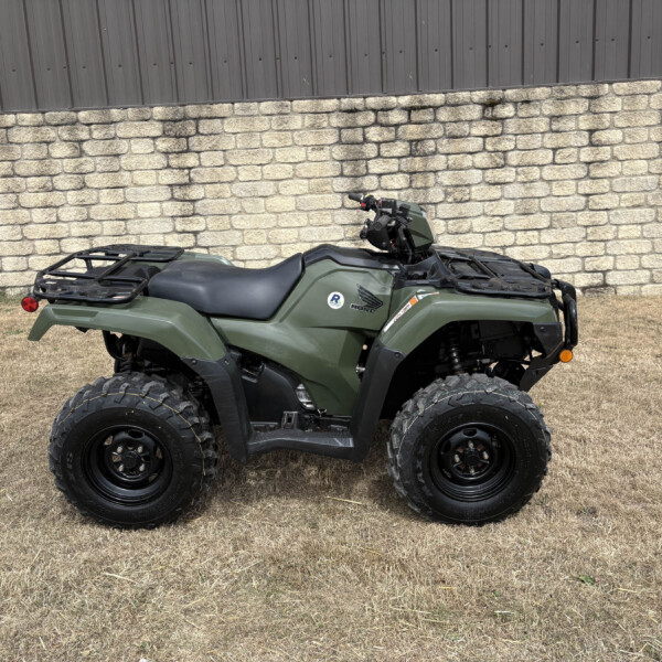Green all-terrain vehicle (ATV) parked on dry grass in front of a stone wall and dark metal siding fence.