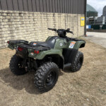 Olive green four-wheel ATV parked on dry grass beside a stone wall and industrial buildings in the background.