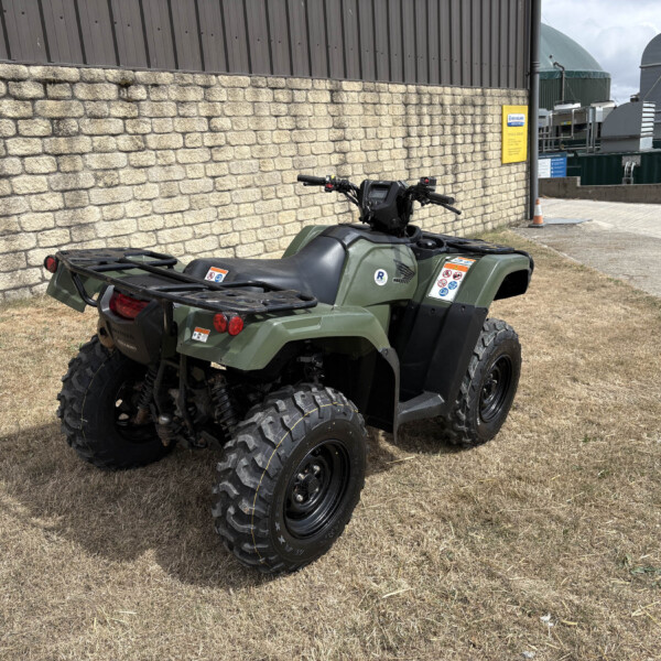 Olive green four-wheel ATV parked on dry grass beside a stone wall and industrial buildings in the background.