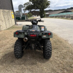Rear view of a green ATV parked on a dry, grassy area along a brick-walled building and industrial sheds.