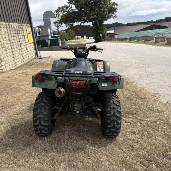 Rear view of a green ATV parked on a dry, grassy area along a brick-walled building and industrial sheds.