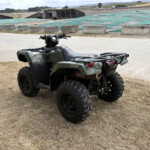 Green Honda ATV with chunky off-road tires parked on a dry dirt lot at a farm, with green tarps and farm buildings in the background.”} ,{