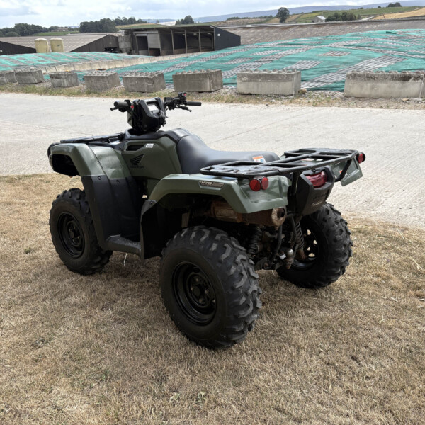 Green Honda ATV with chunky off-road tires parked on a dry dirt lot at a farm, with green tarps and farm buildings in the background.”} ,{